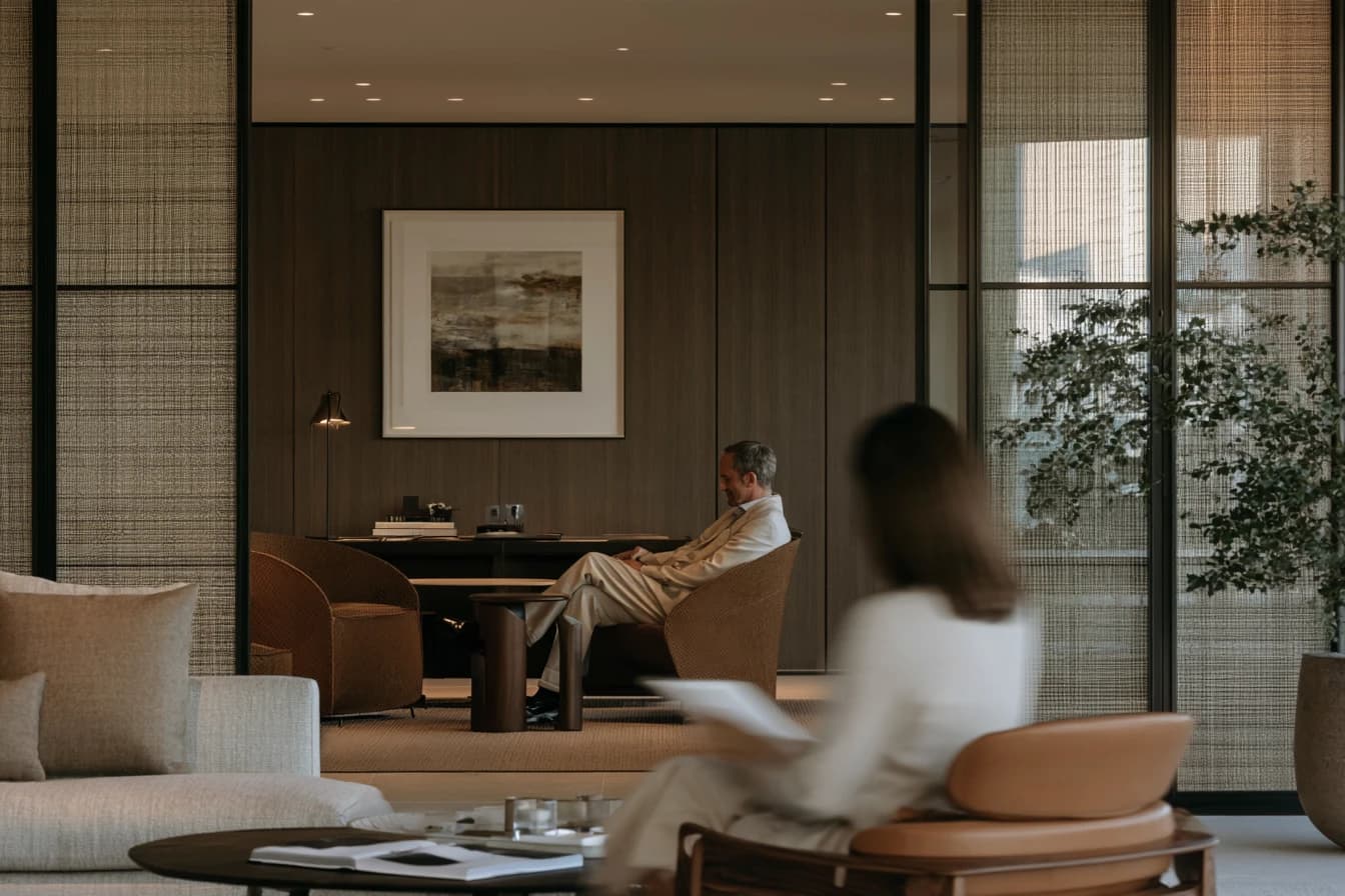 A man in a consulting room seen through glass, a woman reading in the lounge outside