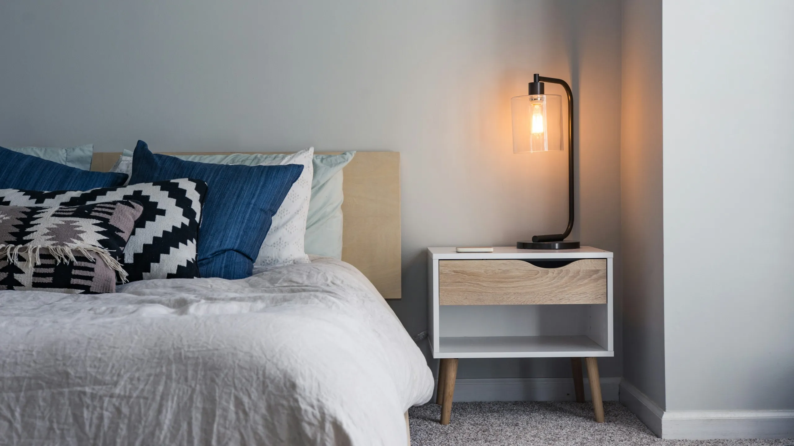 A lamp glowing on a white bedside table beside a made bed with blue cushions