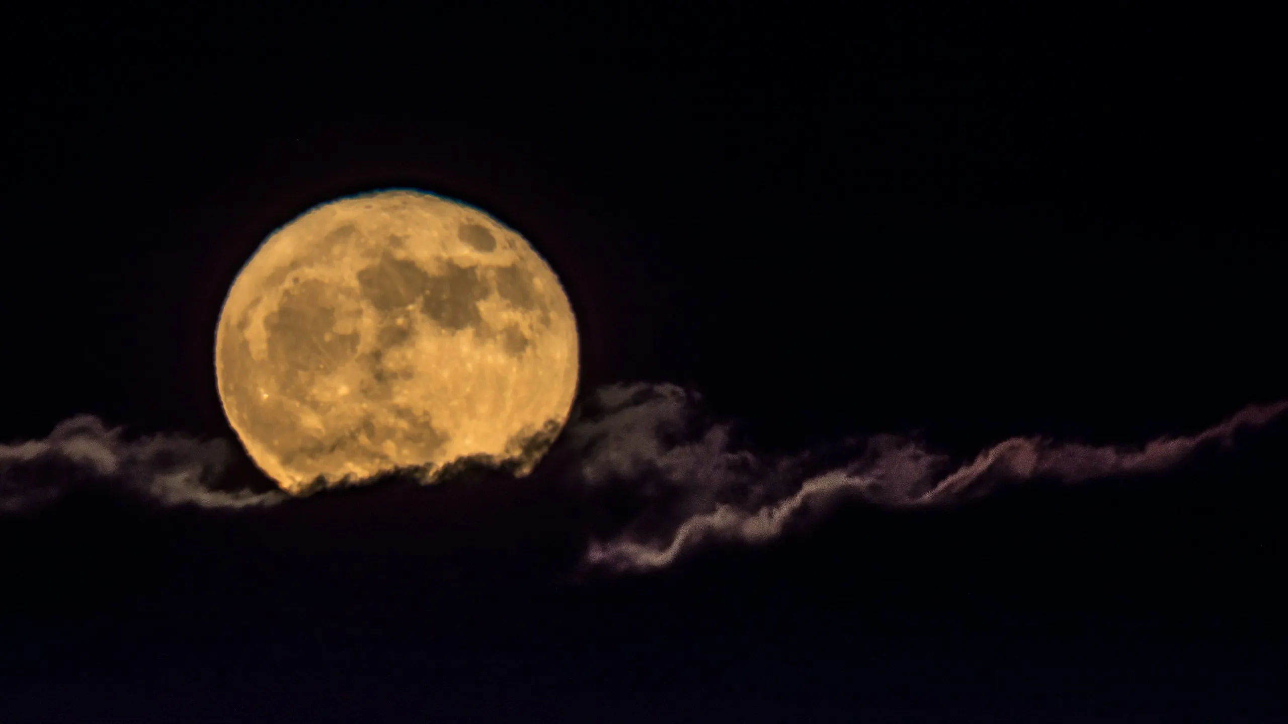 A full moon rising behind a thin band of cloud in a black sky