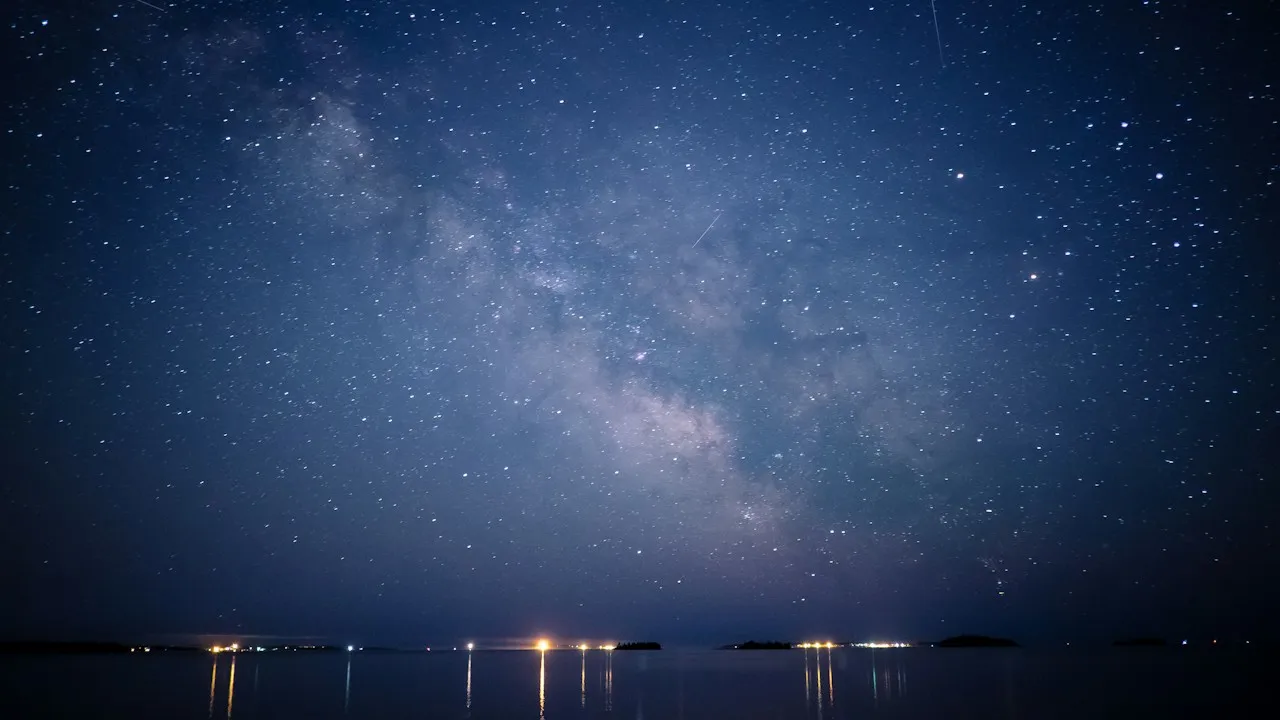 The Milky Way over calm dark water, with distant lights along the shore